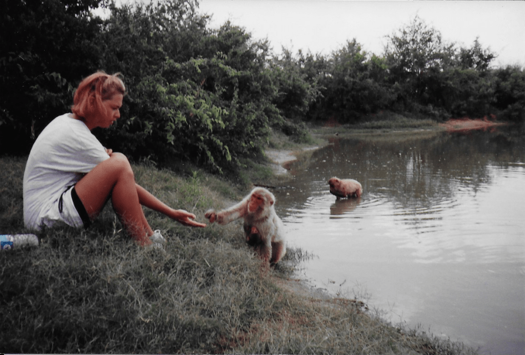Image of women sitting at pond edge reaching hand out with food to a Japanese macaque
